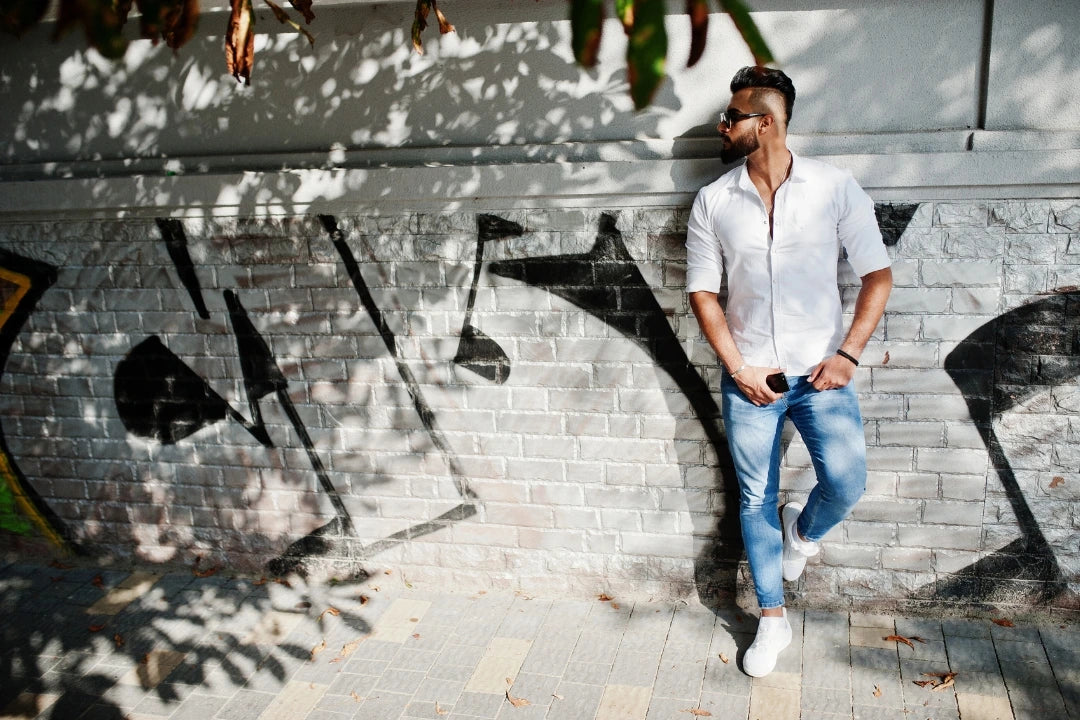 Man in sunglasses wearing a white shirt and blue jeans outdoors beside a graffiti mural.