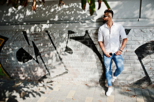Man in sunglasses wearing a white shirt and blue jeans outdoors beside a graffiti mural.