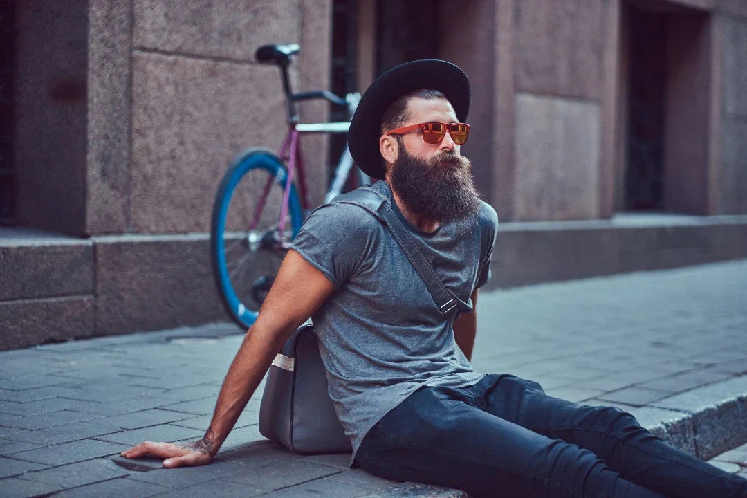 Bearded man wearing a hat and sunglasses sitting on a sidewalk with a bicycle behind him.