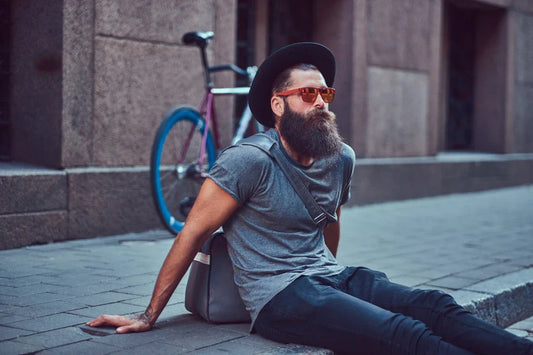 Bearded man wearing a hat and sunglasses sitting on a sidewalk with a bicycle behind him.